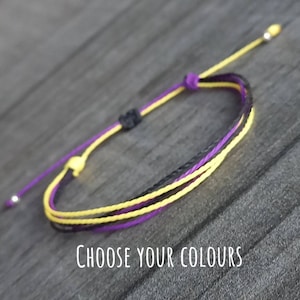 May include: A purple, black and yellow friendship bracelet with silver metal ends. "Choose Your Colours" is written below the bracelet.