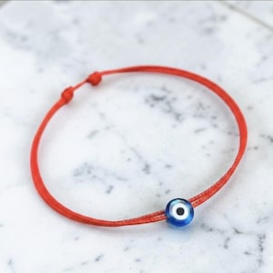 May include: Red string bracelet with a blue and white evil eye bead. The bracelet has an adjustable sliding knot. The evil eye bead is a deep blue with a white and black centre. The bracelet is shown on a marble surface.