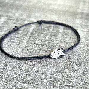 May include: A black cord bracelet with a small, silver-coloured fish charm. The bracelet is adjustable with sliding knots. The fish charm has etched details, including an eye and fins. The bracelet is shown on a grey surface.