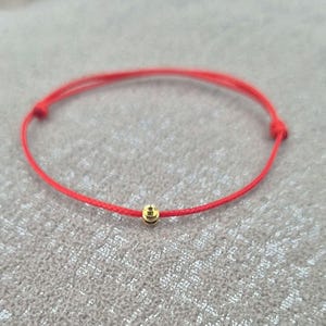 May include: A simple red string bracelet featuring a small gold bead. The bracelet is knotted at each end. The background is a neutral grey.