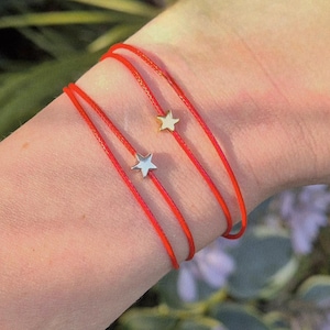 May include: Three red string bracelets, each with a star charm. Two charms are silver, one is gold. The bracelets are worn on a wrist, with a blurred green background.