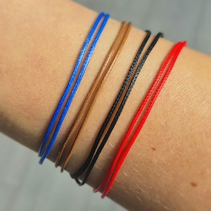 May include: Five thin cord bracelets in blue, brown, black, and red. The bracelets are shown on an arm, highlighting the different colours and textures of the cords. Each bracelet is made of a thin, woven cord.