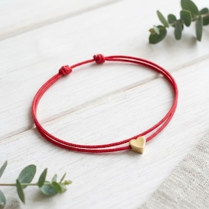 May include: A red cord bracelet featuring a small, smooth, gold-toned heart charm. The bracelet is constructed from a thin, durable red string and includes two adjustable sliding knots to secure the fit around the wrist.