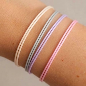 May include: Four thin, double-stranded cord bracelets in pastel colours. The set includes one cream, one light grey, one lavender, and one light pink bracelet, each featuring a smooth, minimalist texture and a simple, continuous circular design.