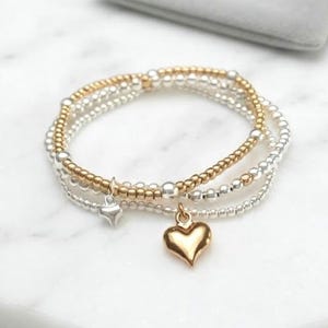 May include: A set of three stacked beaded bracelets featuring small metallic spheres in alternating gold and silver tones. One bracelet includes a polished gold heart charm, while another features a smaller, smooth silver heart-shaped pendant.