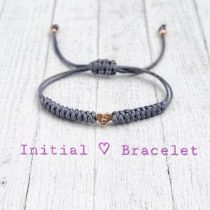 May include: A grey macrame bracelet with a gold heart-shaped charm bearing the letter "B". The bracelet has adjustable sliding knots and rose gold-coloured end caps. The text "Initial Bracelet" is below the bracelet.