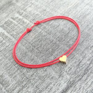 May include: A red string bracelet featuring a small gold heart charm. The bracelet is knotted and has a minimalist design. The heart charm is centrally placed on the bracelet.