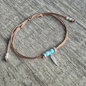 May include: A brown leather cord bracelet with a silver feather charm and two turquoise beads.