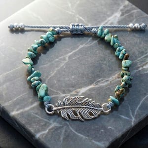 May include: A turquoise stone bracelet with a silver feather charm. The bracelet features irregular turquoise stones strung on a grey cord, with a sliding knot closure. The feather charm is detailed and adds an elegant touch.