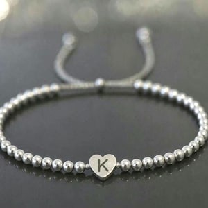 May include: Silver beaded bracelet with a heart-shaped charm. The charm features the letter "K" in the centre. The bracelet has an adjustable sliding knot closure. The beads are small and round.