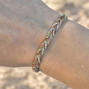 May include: A braided bracelet with silver, gold, and copper-coloured threads. The bracelet is displayed on a wrist, highlighting the detailed weave and metallic shades. This accessory offers a mix of colours and textures.