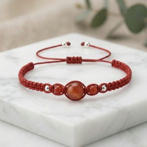 May include: A red macrame cord bracelet featuring a central round orange-red gemstone bead, flanked by two smaller matching beads and two silver-toned metal beads. The bracelet includes an adjustable sliding knot closure with two silver-toned end beads.