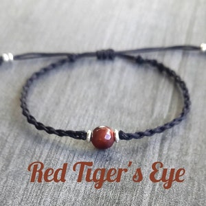 May include: A black braided cord bracelet with a single red tiger's eye bead. The bracelet is tied with a slip knot.