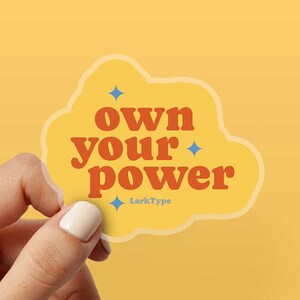 Può includere: Un adesivo giallo con le parole "own your power" in arancione, con accenti di stelle blu. L'adesivo ha una forma a nuvola e il testo "LarkType" in basso. L'adesivo è tenuto da una mano su uno sfondo giallo.