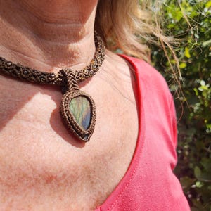 May include: A handmade macramé necklace with a teardrop-shaped pendant containing an iridescent stone. The necklace is made with brown cord, and the pendant has blues, greens, and yellows. The necklace is worn by a person wearing a red top.