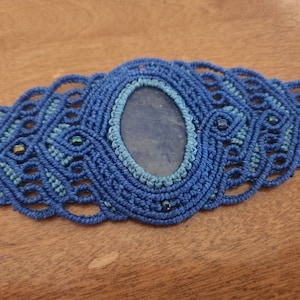 Puede incluir: Un brazalete de macramé azul con una gran piedra preciosa azul en el centro. El brazalete está decorado con pequeñas cuentas azules.