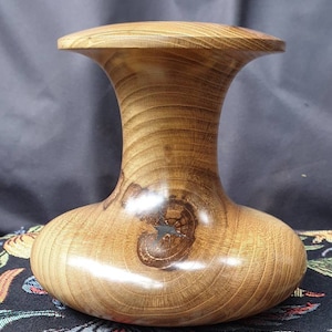 May include: A wooden vase with a wide, rounded base and a narrow neck. The vase is made of light brown wood with a dark brown knot in the center. The vase is polished and has a smooth finish.