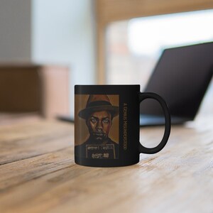Pu&ograve; includere: Tazza in ceramica nera con un ritratto di un uomo con un cappello marrone e una camicia bianca. La tazza ha il testo "A GENIUS MISUNDERSTOOD" stampato sul lato.