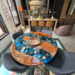 May include: A round wooden table with a blue epoxy resin top. The table is decorated with small bowls filled with various items, including nuts, dried fruit, and small bottles. The table is surrounded by four chairs with grey fabric seats.