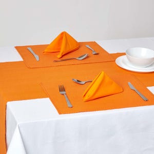 May include: Two orange placemats with folded orange napkins and silverware on a white tablecloth.