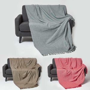 May include: Three different coloured throw blankets draped over grey sofas. The blankets are a light grey, a brown, and a red. Each blanket has a fringe border.