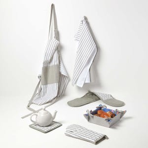 May include: A selection of kitchen items in a neutral colour scheme. The image shows a striped apron, tea towels, oven mitt, pot holders, a teapot, and a bread basket with a floral lining. The items are grey and white.