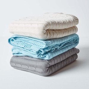 May include: A stack of four cable knit throw blankets in different colours. The top blanket is cream, the second is light blue, the third is grey, and the bottom is a darker grey.