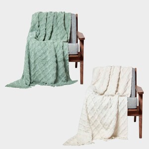 May include: Two throw blankets, one green and one white, are draped over wooden chairs with grey cushions. The blankets have a cable knit pattern and fringe.