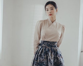 May include: A woman wearing a beige blouse and a long, floral skirt. The skirt has a dark blue background with white and pink flowers. She is wearing black ankle boots with white embellishments.