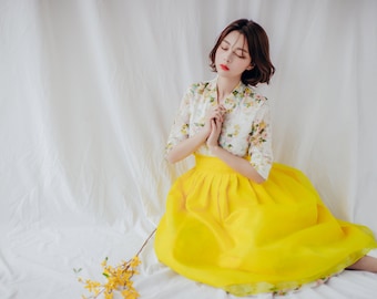 May include: A woman wearing a traditional Korean Hanbok. The top is white with a floral pattern, and the skirt is a bright yellow. The outfit is paired with white shoes. A branch of yellow flowers lies nearby.
