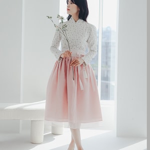 Könnte beinhalten: Eine Frau trägt eine weiße Bluse mit Blumenmuster und einen rosa Rock. Sie hält einen Zweig mit weißen Blüten.