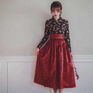 May include: A woman wearing a traditional Korean hanbok dress. The dress is black with floral print and a red skirt. She is standing in front of a white wall with a light fixture above her head.
