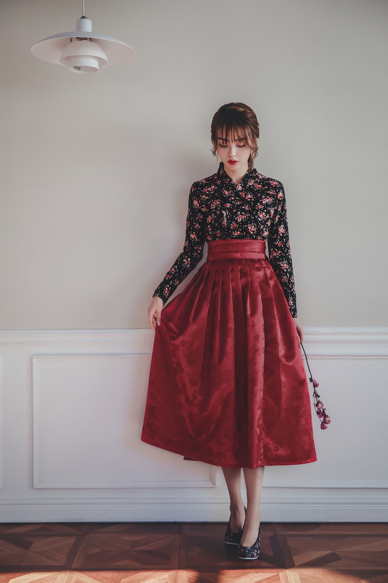 May include: A woman wearing a traditional Korean hanbok dress. The dress is black with floral print and a red skirt. She is standing in front of a white wall with a light fixture above her head.
