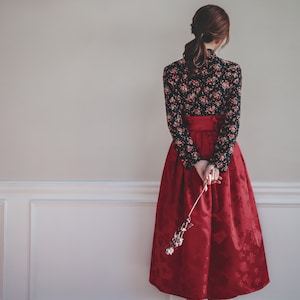 May include: A woman wearing a red skirt with a black floral print top and black heels. She is holding a dried flower in her hand.