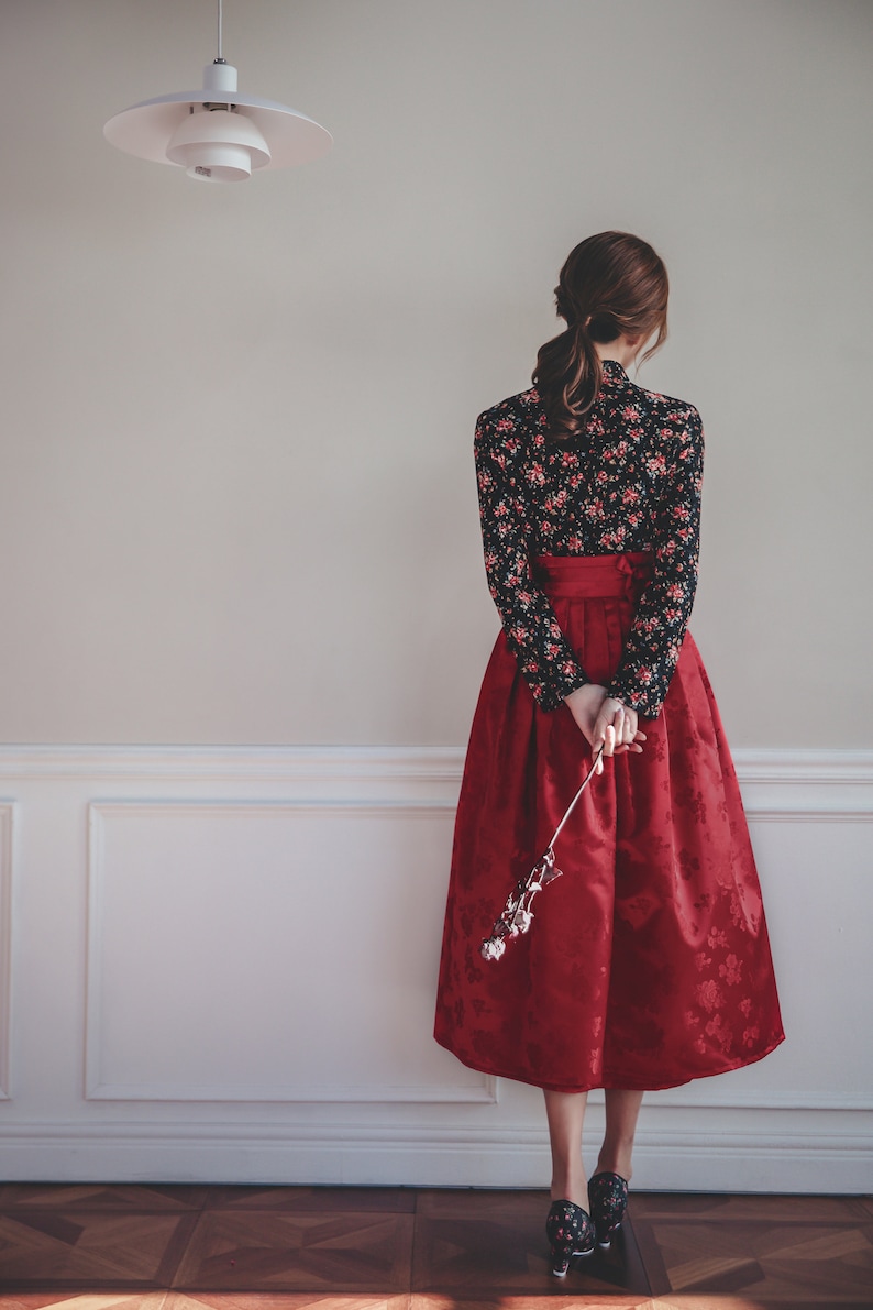 May include: A woman wearing a red skirt with a black floral print top and black heels. She is holding a dried flower in her hand.