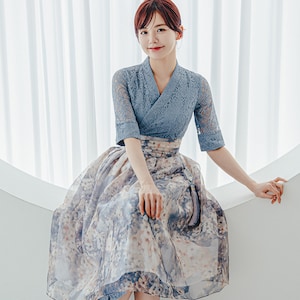 May include: A woman wearing a traditional Korean Hanbok dress. The top is a light blue lace with three-quarter sleeves and a V-neck. The skirt is a flowing floral print in shades of blue, beige, and pink.