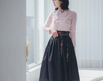 May include: A woman wearing a traditional Korean hanbok dress. The hanbok is a light pink top with a black skirt and a floral patterned sash. The woman is standing in a white room with a window in the background.