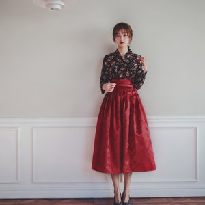 May include: A woman wearing a black floral blouse and a red skirt with a pleated design. She is standing in front of a white wall with white trim.