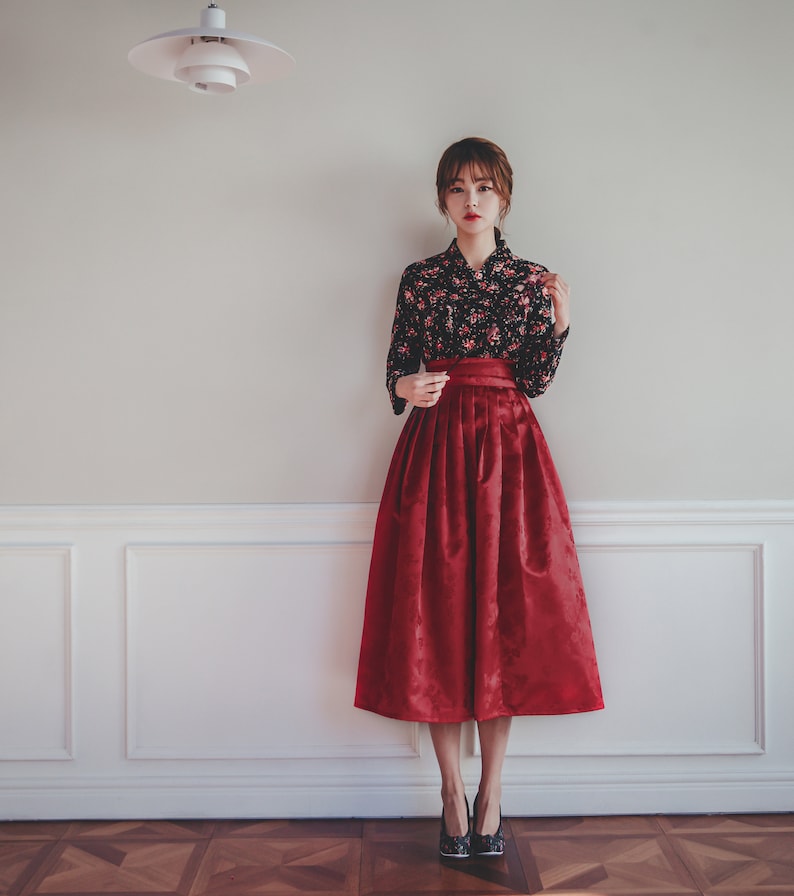 May include: A woman wearing a black floral blouse and a red skirt with a pleated design. She is standing in front of a white wall with white trim.