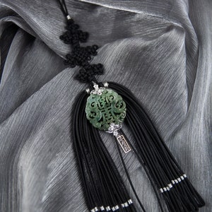 Puede incluir: Un colgante de jade verde y negro con una borla que cuelga de una cuerda negra. El colgante tiene caracteres chinos tallados en él. La borla está hecha de muchos hilos de cuerda negra.