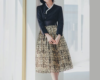 May include: A woman wearing a black hanbok with a white collar and a black sash. The skirt is a light yellow with a black floral pattern.