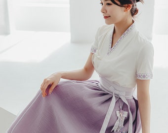 May include: A traditional Korean hanbok ensemble. The outfit features a white short-sleeved top with purple floral trim and a V-neck. The skirt is a soft lavender color with a matching sash and decorative embellishments.