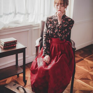 May include: A woman wearing a traditional Korean hanbok dress with a black floral top and a red skirt. She is sitting in a chair with her hands clasped in her lap. The chair is in front of a window with white curtains.