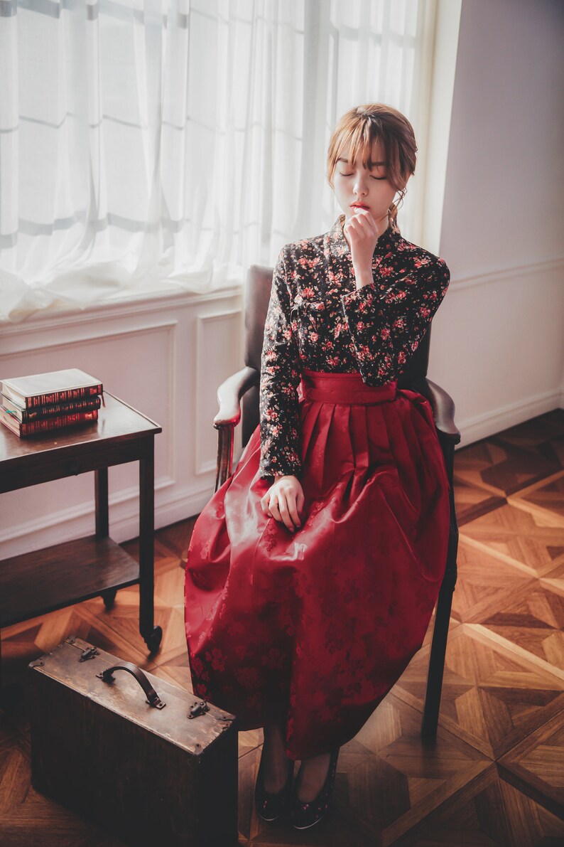 May include: A woman wearing a traditional Korean hanbok dress with a black floral top and a red skirt. She is sitting in a chair with her hands clasped in her lap. The chair is in front of a window with white curtains.
