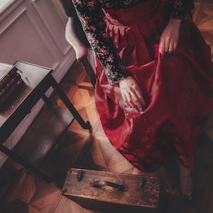 May include: A woman wearing a red and black dress sits on a chair in a room with wooden floors. A small wooden table with a stack of books sits in the foreground.