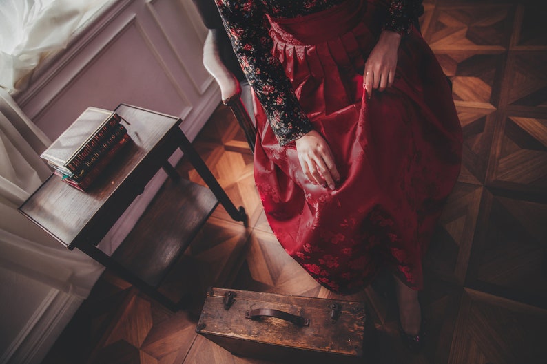 May include: A woman wearing a red and black dress sits on a chair in a room with wooden floors. A small wooden table with a stack of books sits in the foreground.