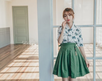 May include: A woman wearing a white blouse with a floral pattern and a green skirt with a gold tassel. She is standing in a room with a white door and a window.