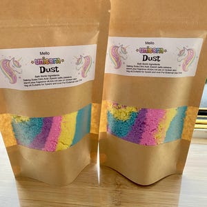 May include: Two brown paper pouches with clear windows, filled with colourful bath bomb powder. Each pouch has a label with a unicorn graphic and the words "Unicorn Dust". The powder is a mix of blue, yellow, pink, and purple.