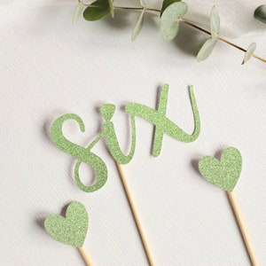Può includere: Topper per torta di glitter verde che compongono la parola "sei" con due topper a forma di cuore di glitter verde.