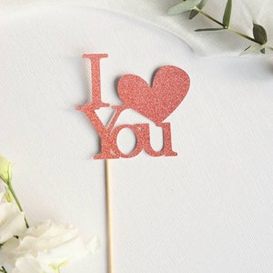 Puede incluir: Un adorno para pastel de purpurina rosa con las palabras "Te amo" y un corazón.